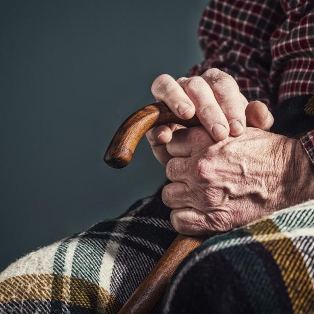 Eine ältere Person hält einen Holzstock in ihren Händen, bedeckt mit einer karierten Decke.