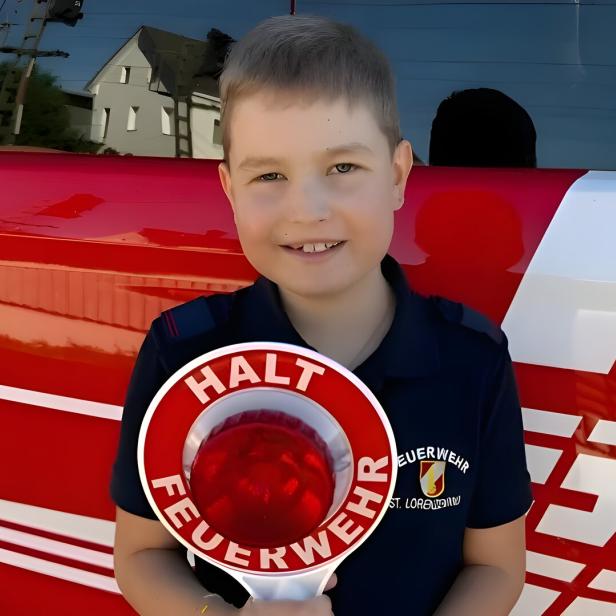 Ein Junge in Feuerwehrkleidung hält ein Stoppschild vor einem roten Feuerwehrfahrzeug.