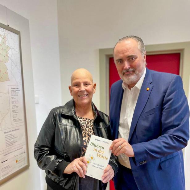 Zwei Personen stehen nebeneinander und halten gemeinsam ein Buch mit dem Titel „Blumen in der Wüste“.
