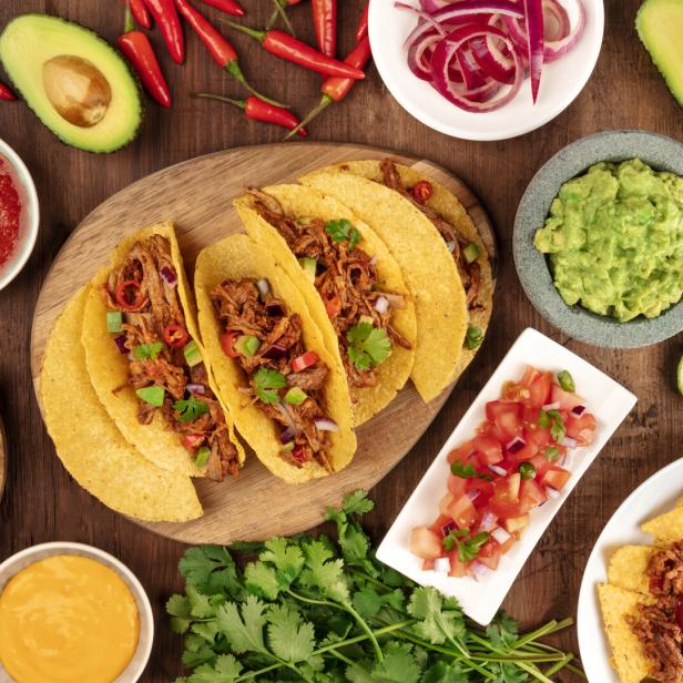 Mexikanisches Essen mit Tacos, Tortilla-Chips, Guacamole, Salsa, Käse, Limetten, Chili und Jalapeños auf Holztisch.