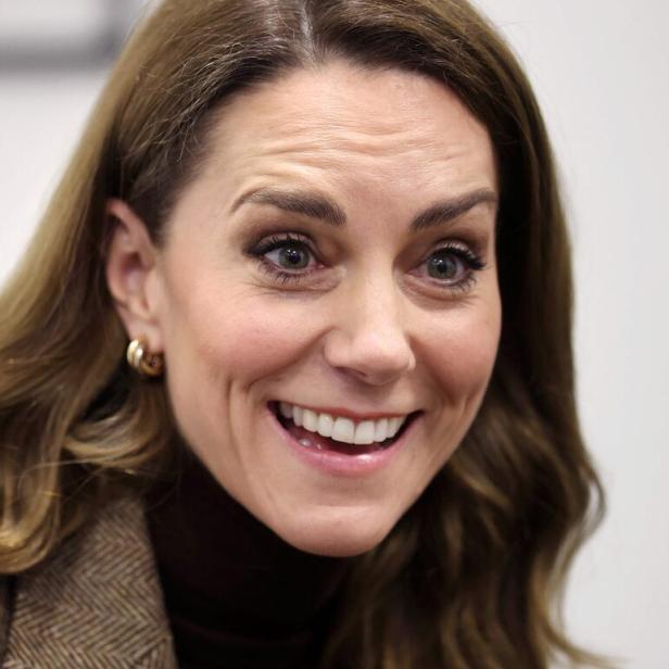 Prinzessin Kate mit braunen Haaren und braunen Augen lächelt freundlich, trägt braunen Rollkragenpullover und Ohrringe.