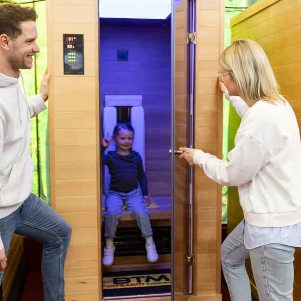Ein Kind sitzt in einer Infrarotkabine, zwei Erwachsene stehen lächelnd davor.
