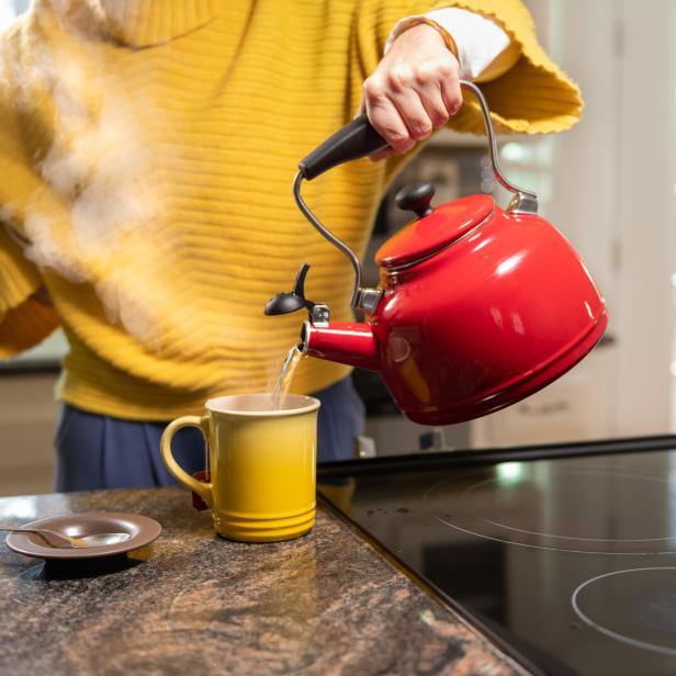 Eine Person gießt heißes Wasser aus einem roten Wasserkessel in eine gelbe Tasse auf einer Küchenarbeitsplatte.