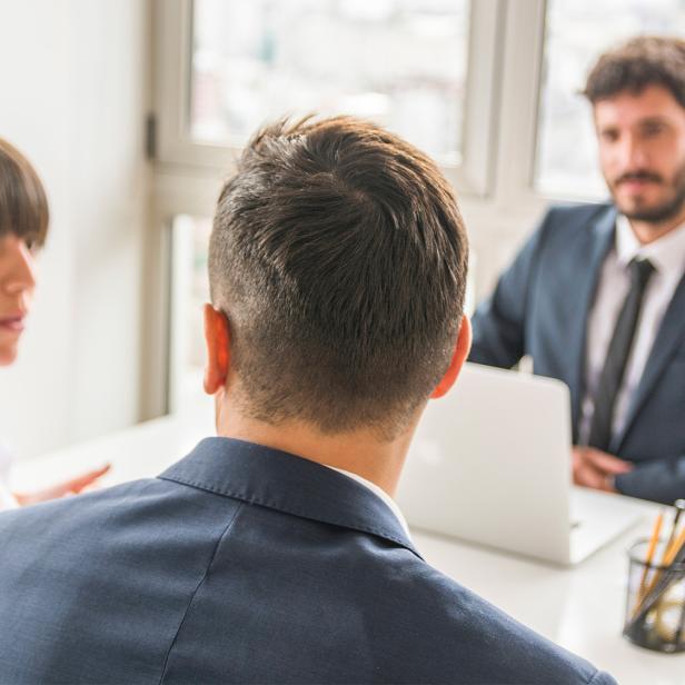 Zwei Männer und eine Frau sitzen in Business-Klamotten in einem lichtdurchfluteten Büroraum. 