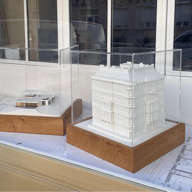 Zwei Architekturmodelle in Vitrinen auf Holzsockeln, ausgestellt auf Bauplänen vor einem Fenster.