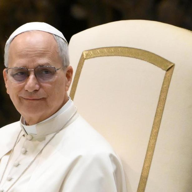 Ein Mann mit Brille und weißem Gewand sitzt auf einem verzierten Stuhl.
