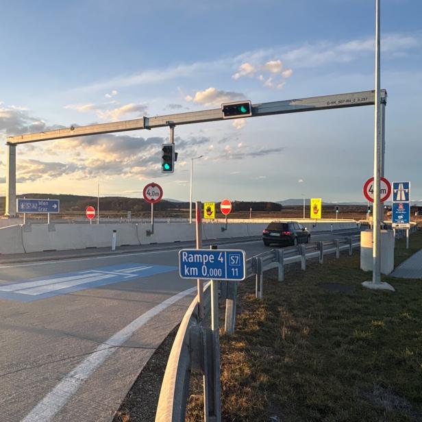 Eine Autobahnauffahrt mit Verkehrszeichen, Ampeln und einem Auto ist zu sehen.