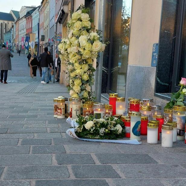 Kerzen, Blumen und ein Kreuz aus weißen Rosen stehen als Gedenkstätte am Straßenrand einer belebten Fußgängerzone.
