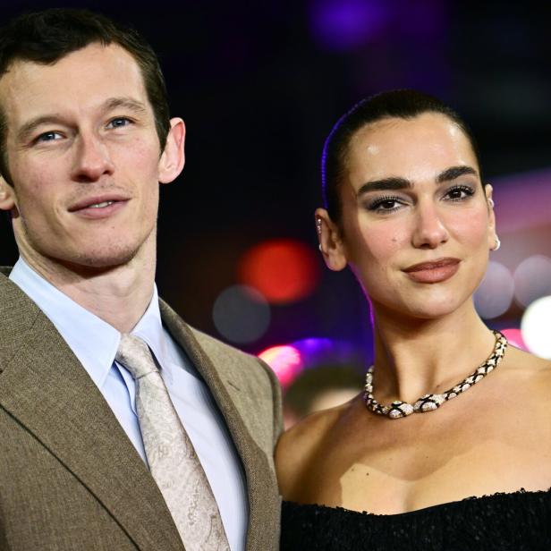 Callum Turner und seine Partnerin, Sängerin Dua Lipa mit Halskette und schulterfreiem Kleid, stehen nebeneinander vor bunten Lichtern.