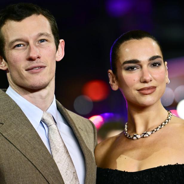 Callum Turner und seine Partnerin, Sängerin Dua Lipa mit Halskette und schulterfreiem Kleid, stehen nebeneinander vor bunten Lichtern.