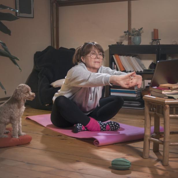 Eine Frau macht Dehnübungen auf einer Yogamatte vor einem Laptop, während ein kleiner Hund auf einem Kissen sitzt.