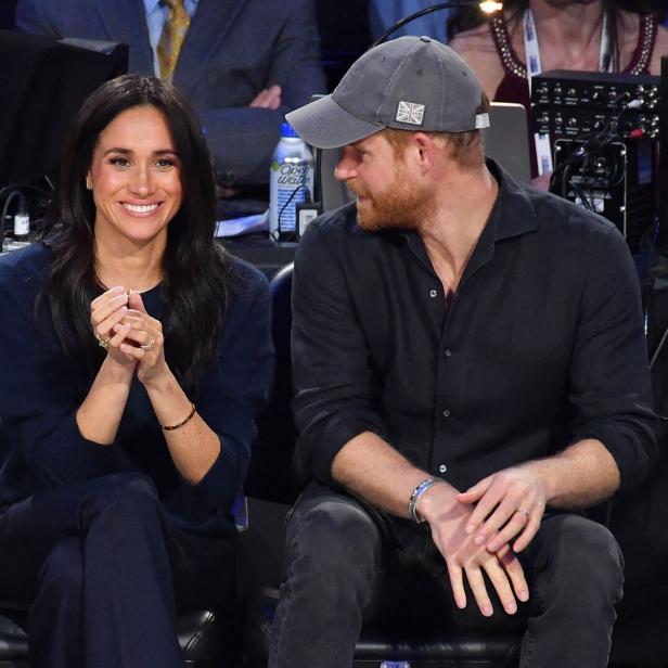 Meghan und Harry sitzen nebeneinander, sie lächelt, er trägt eine Kappe.