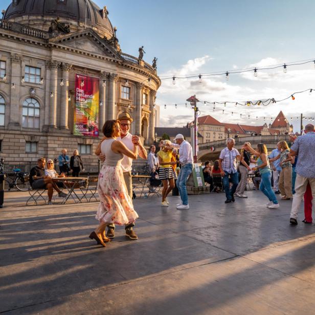 Mehrere Menschen tanzen fröhlich im Freien vor einem historischen Gebäude bei Sonnenuntergang.