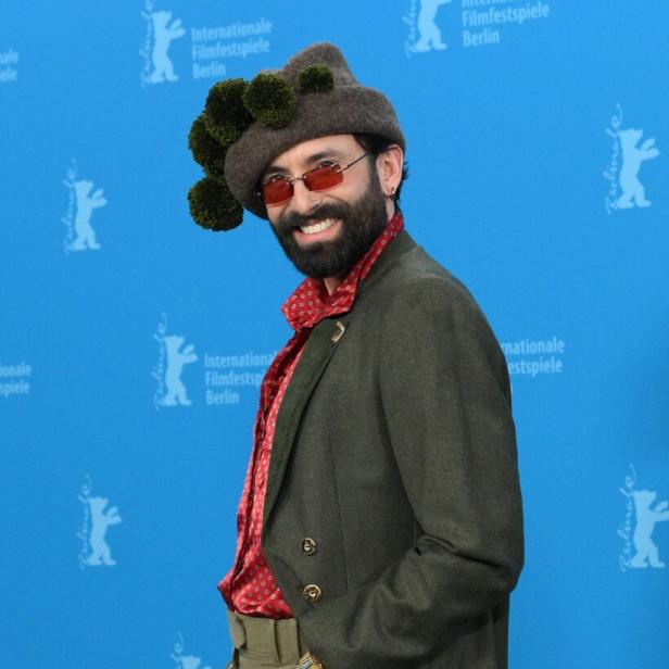 Eine Person mit auffälligem Hut, rotem Hemd, grünem Sakko und Sonnenbrille posiert vor blauem Berlinale-Hintergrund.