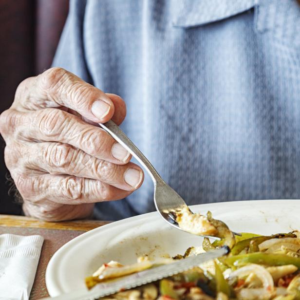 Eine ältere Hand hält eine Gabel über einem Teller mit gebratenem Gemüse und Fleisch.