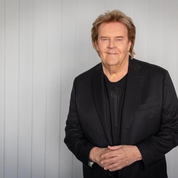 Howard Carpendale mit blondem Haar und schwarzem Sakko lächelt vor einem hellen Hintergrund.