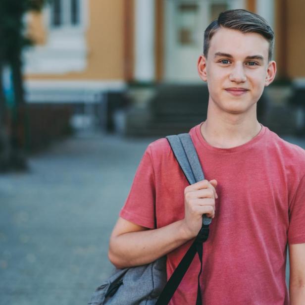 Jugendlicher mit Rucksack steht draußen vor einem Gebäude und lächelt in die Kamera.