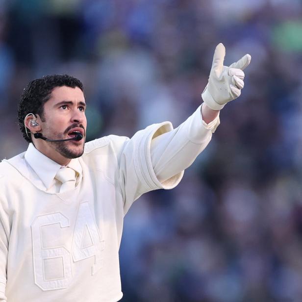 Musiker Bad Bunny mit Headset und weißen Handschuhen trägt ein weißes Oberteil mit der Zahl 64 und zeigt nach oben.