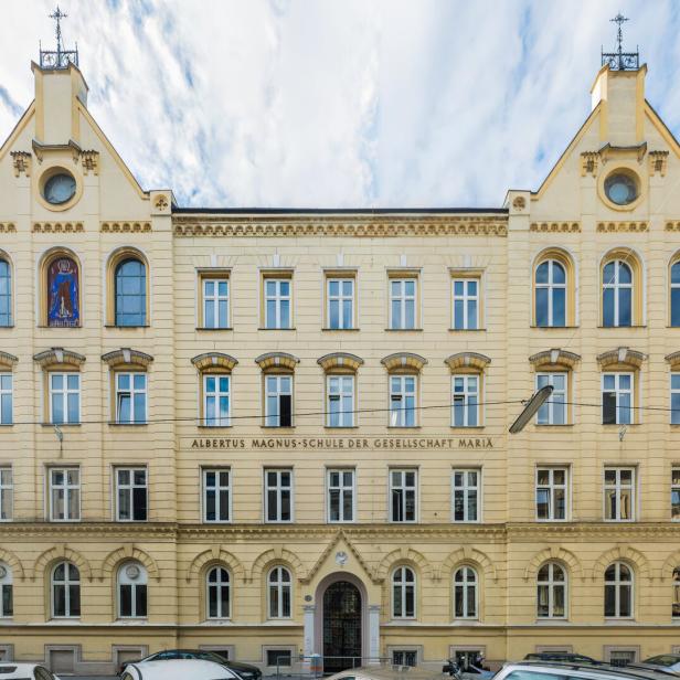 Albertus Magnus Gymnasium: Historisches, gelbes Schulgebäude mit symmetrischer Fassade, vielen Fenstern und zwei Giebeln, davor parken Autos.