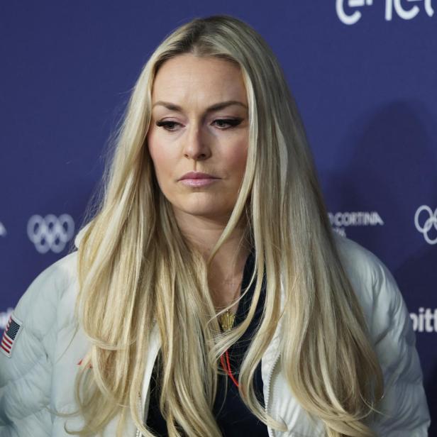 Lindsey Vonn mit blonden Haaren und weißer Jacke vor blauem Hintergrund mit Olympia-Logo.