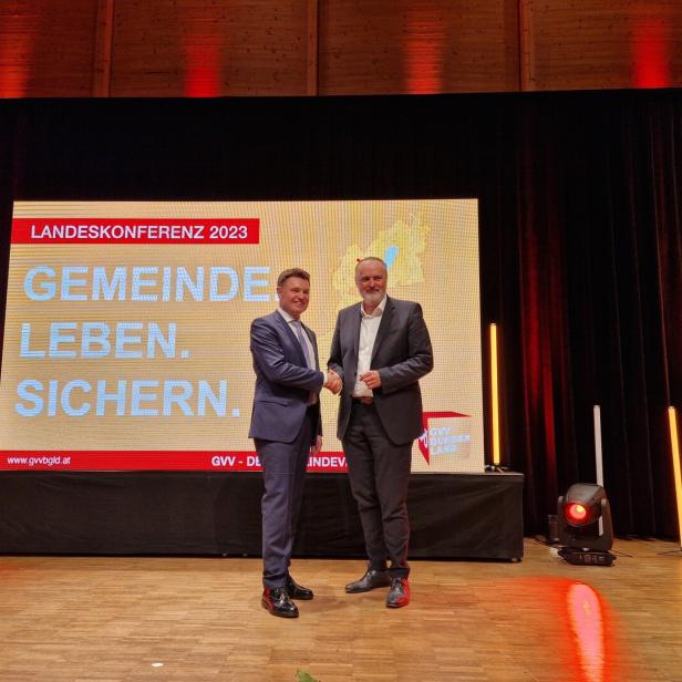 Zwei Männer im Anzug geben sich auf einer Bühne vor einem großen Bildschirm mit Konferenzschriftzug die Hand.