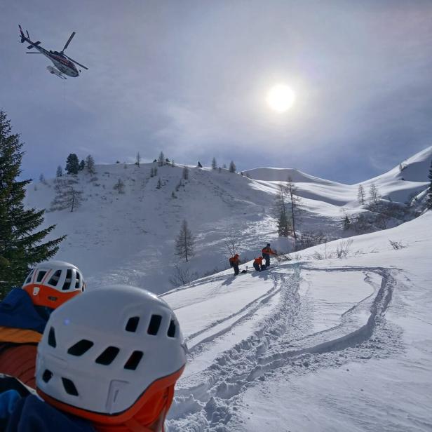 Ein Hubschrauber fliegt über eine verschneite Berglandschaft, während Bergretter zu sehen sind.