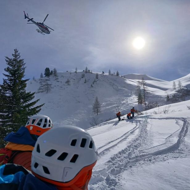 Ein Hubschrauber fliegt über eine verschneite Berglandschaft, während Bergretter zu sehen sind.