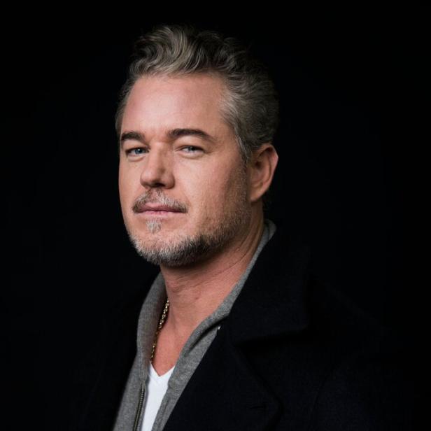 Eric Dane mit grau meliertem Haar und Bart trägt einen dunklen Mantel über einem grauen Hoodie und einem weißen T-Shirt.