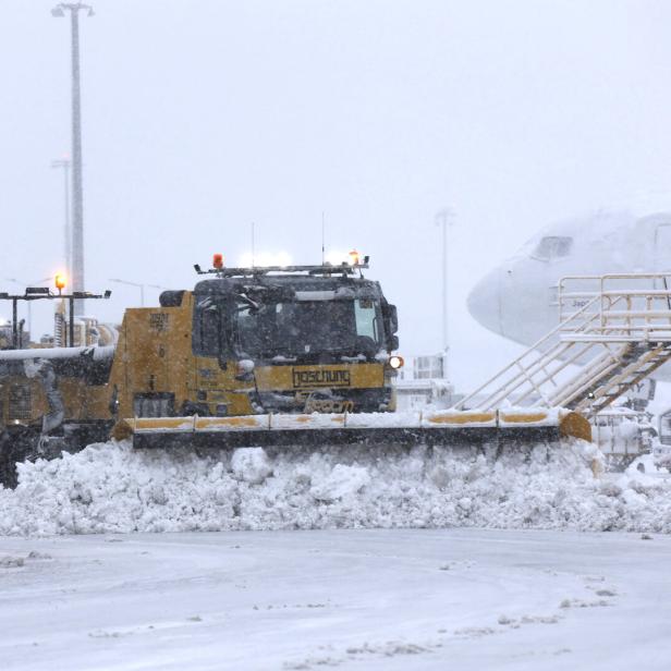 Ein gelber Schneepflug räumt Schnee auf einem Flughafen, im Hintergrund ein Flugzeug.