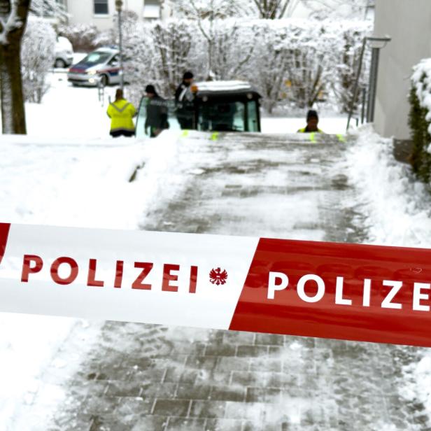 Ein rot-weißes Polizei-Absperrband versperrt einen verschneiten Weg, im Hintergrund sind Einsatzkräfte und ein Polizeiauto zu sehen.
