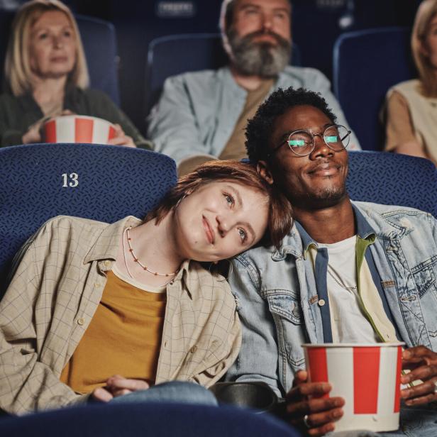 Ein junges Paar sitzt eng aneinandergelehnt im Kino und genießt Popcorn, umgeben von weiteren Zuschauern.