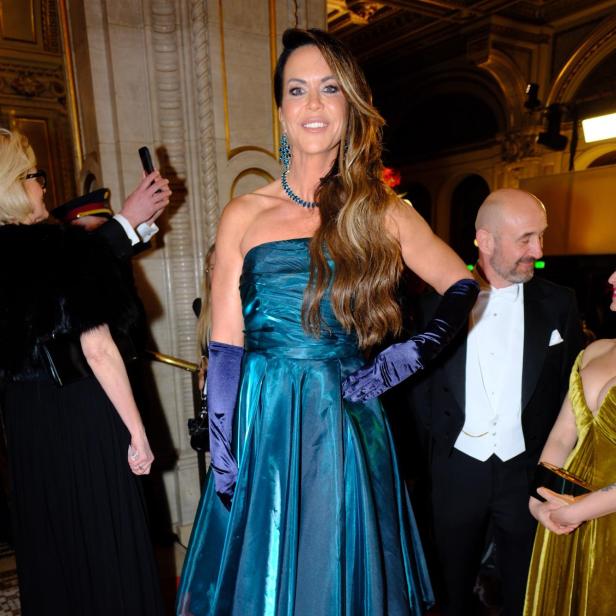 Frau in schimmerndem türkisfarbenem Abendkleid mit langen Handschuhen und Schmuck auf rotem Teppich.