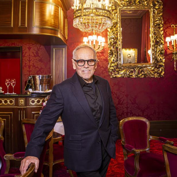 Wolfgang Puck mit Brille und dunklem Anzug steht in einem opulenten, rot dekorierten Raum mit goldenem Spiegel.