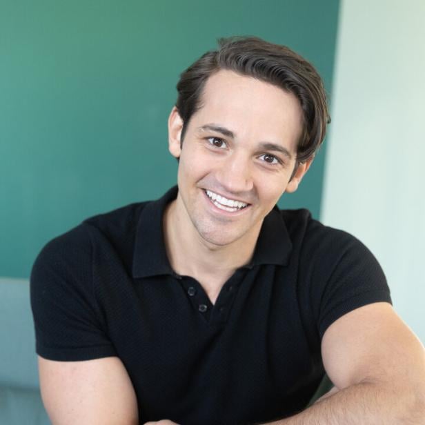 Ein junger Mann mit dunklem Haar und schwarzem Poloshirt sitzt lächelnd auf einem Stuhl und trägt eine weiße Armbanduhr.