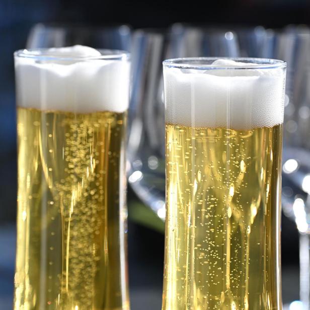 Zwei Gläser mit hellem Bier und Schaum stehen im Vordergrund, im Hintergrund unscharfe Weingläser.