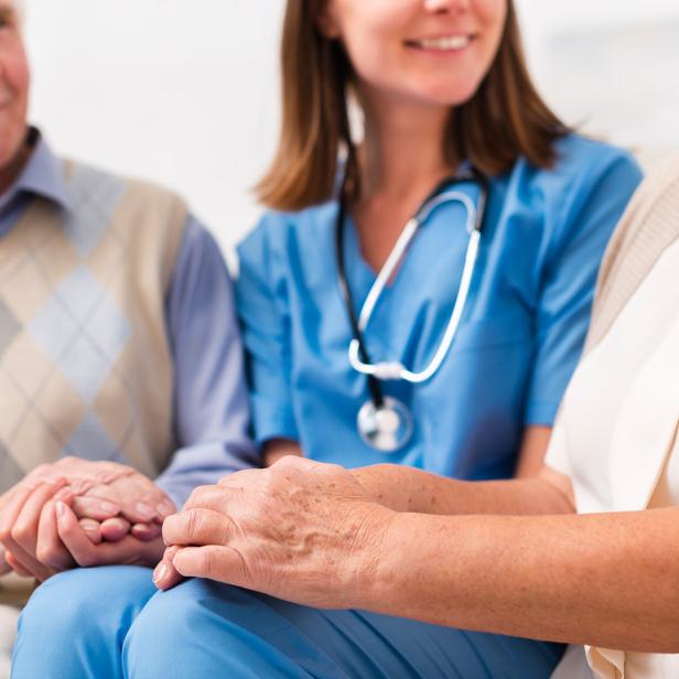 Pflegekraft im Gespräch mit älterem Paar, alle lächeln und halten sich an den Händen.