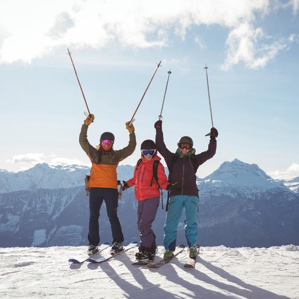 Drei Skifahrer stehen auf einer verschneiten Bergspitze und heben ihre Skistöcke in die Luft.