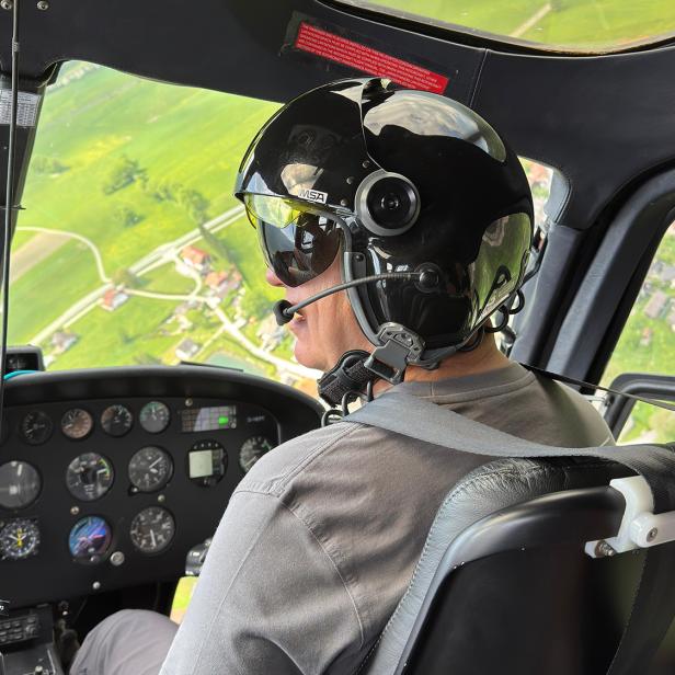 Pilot mit Helm steuert einen Helikopter über eine grüne Landschaft in Österreich.