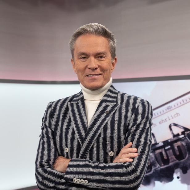Ein Mann mit grau melierten Haaren trägt einen gestreiften Blazer und einen weißen Rollkragenpullover und steht mit verschränkten Armen.