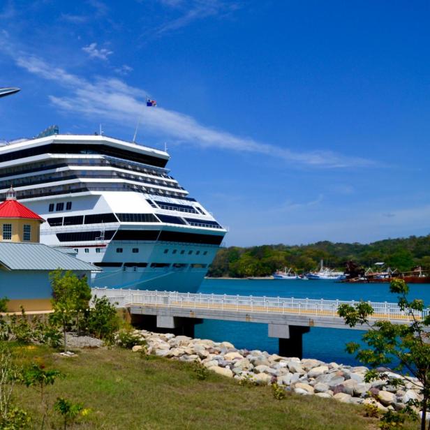Ein großes Kreuzfahrtschiff liegt an einem sonnigen Hafen, während Passagiere durch eine bunte Hafenanlage mit kleinen Gebäuden an Land gehen.