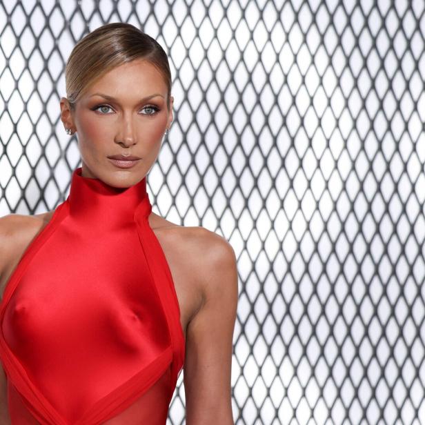 Bella Hadid mit blonden Haaren trägt ein rotes Kleid vor einem Netzgitter.