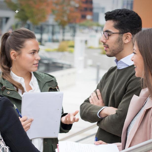 Vier junge Menschen unterhalten sich mit Notizblöcken auf dem Campus der WU Wien.