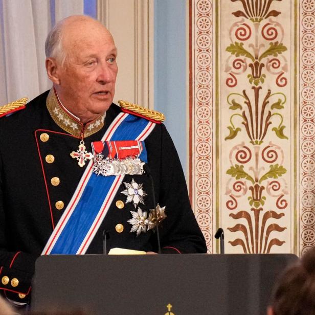 König Harald V. in Uniform mit Orden und blauem Band vor gemusterter Wand.