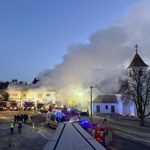 Dichter Rauch steigt aus einem brennenden Gebäude neben einer Kirche auf, zahlreiche Einsatzfahrzeuge und Feuerwehrleute sind vor Ort.