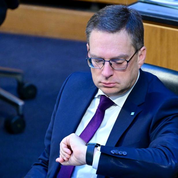 Ein Mann im Anzug mit Brille sitzt auf einem Stuhl und schaut auf seine Armbanduhr.