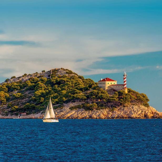 Eine kleine Insel mit grünem Bewuchs, einem rot-weißen Leuchtturm, einem Haus und einem Segelboot im Vordergrund.