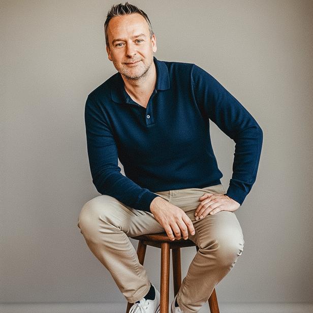 Mann in dunkelblauem Pullover sitzt entspannt auf einem Hocker vor neutralem Hintergrund.