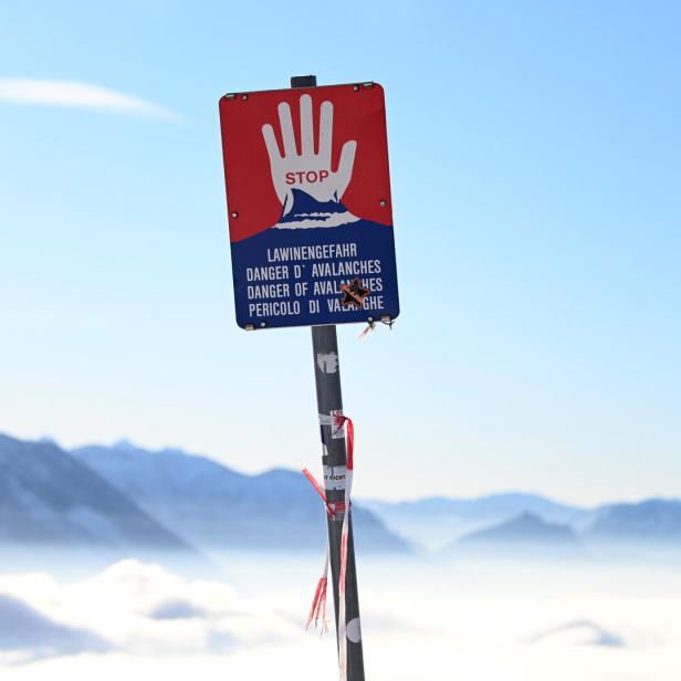 Warnschild mit roter Hand und Hinweis auf Lawinengefahr vor verschneiten Bergen und blauem Himmel.