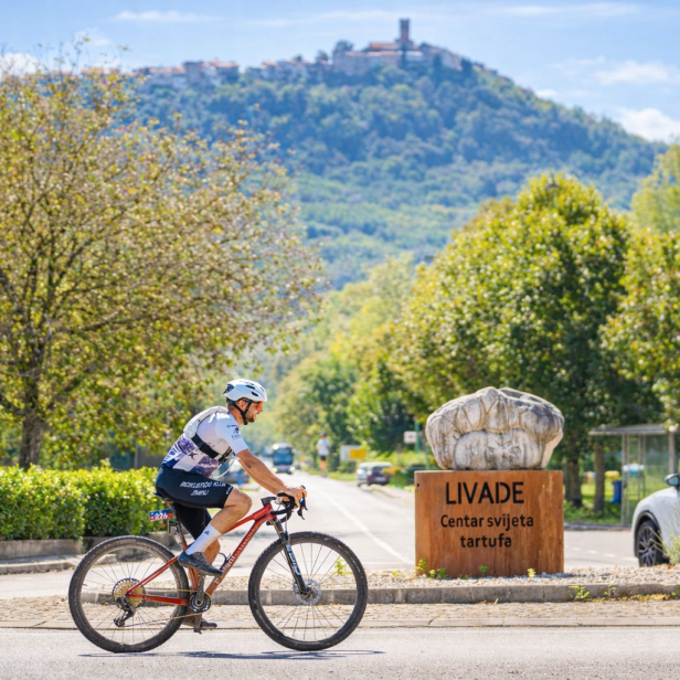 Radfahrer fahren an einem Schild für Livade vorbei, im Hintergrund ein Hügel mit Stadt.