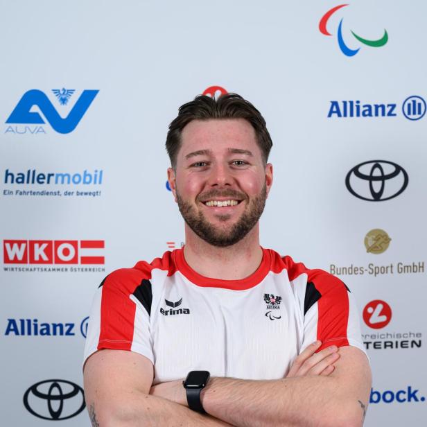 Ein Mann mit verschränkten Armen trägt ein rot-weißes Sporttrikot und lächelt vor einer Sponsorenwand.